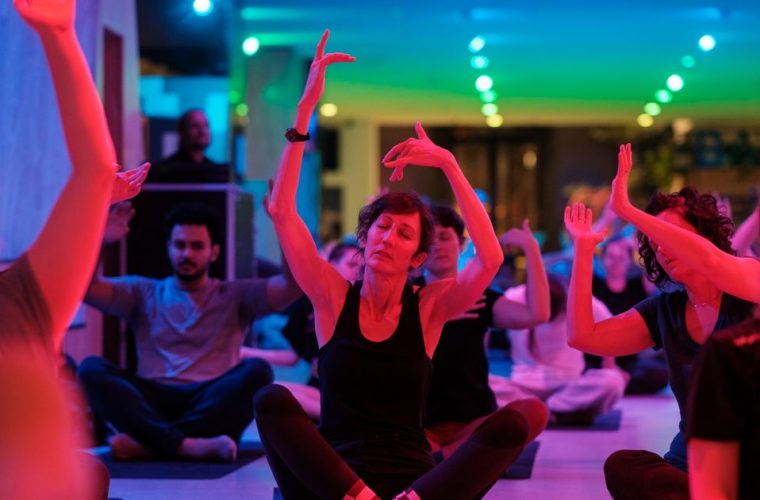 Deelnemers voeren yogabewegingen uit op matten onder de open lucht, ondersteund door techno-muziek